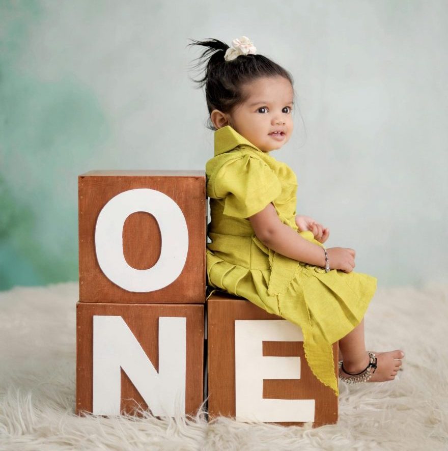 Cute toddler in yellow dress posing with 'ONE' blocks for first birthday photo.